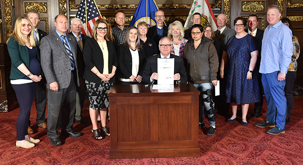 Gov. Tim Walz seated at a desk holding a copy of the signed bill, as the other people in the picture gather around.